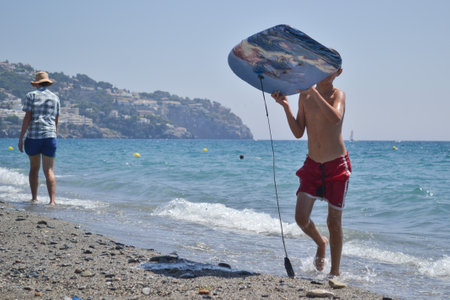 Boy coming out of the sea with surfboard surrounded by other bathers and surfboards with sails in the seaの写真素材