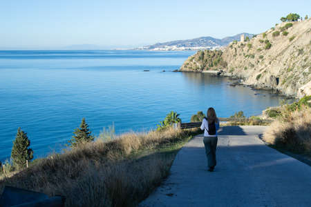 woman with blonde hair going down a small road to the beach of CaÃ±uelo of Nerja with a watchtower and the town of Nerja in the background. Maro Cerro Gordo Cliffsの写真素材