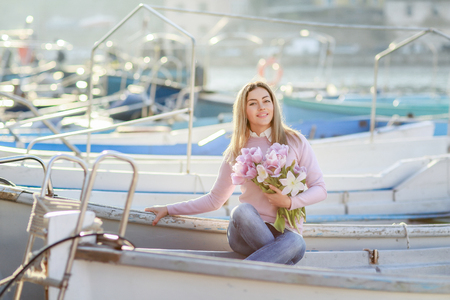 Beautiful girl in spring or autumn, by the sea in the boat Bay. With a bouquet of flowers in his hands and tight stylish jeans and a pink sweater.の写真素材