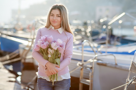 Beautiful girl in spring or autumn, by the sea in the boat Bay. With a bouquet of flowers in his hands and tight stylish jeans and a pink sweater.の写真素材