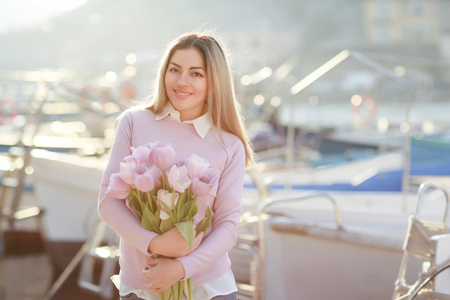 Beautiful girl in spring or autumn, by the sea in the boat Bay. With a bouquet of flowers in his hands and tight stylish jeans and a pink sweater.の写真素材