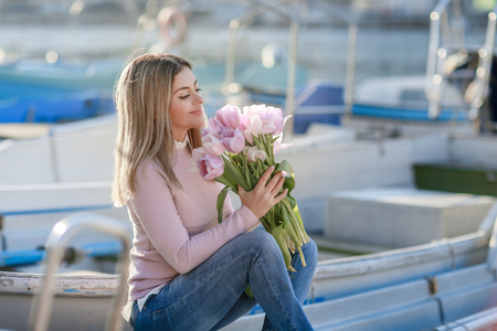 Beautiful girl in spring or autumn, by the sea in the boat Bay. With a bouquet of flowers in his hands and tight stylish jeans and a pink sweater.の写真素材