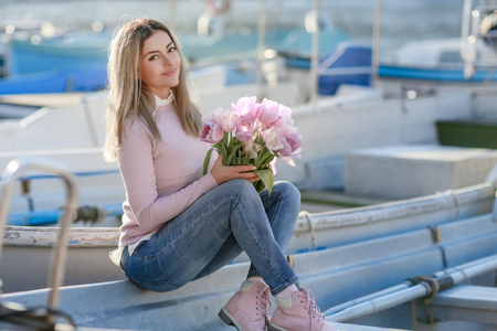 Beautiful girl in spring or autumn, by the sea in the boat Bay. With a bouquet of flowers in his hands and tight stylish jeans and a pink sweater.の写真素材