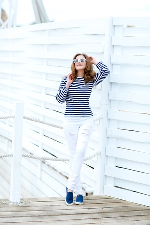 A girl in white tight jeans and sports shoes posing against a white wooden wall. In a sailor t-shirt and glasses. Curly long hair natural color.の写真素材