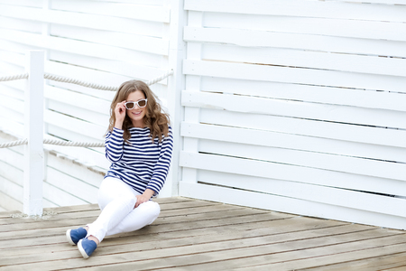 A girl in white tight jeans and sports shoes posing against a white wooden wall. In a sailor t-shirt and glasses. Curly long hair natural color.の写真素材
