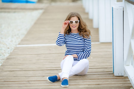 A girl in white tight jeans and sports shoes posing against a white wooden wall. In a sailor t-shirt and glasses. Curly long hair natural color.の写真素材