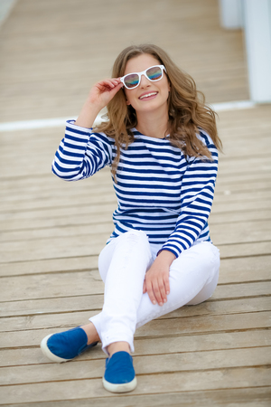 A girl in white tight jeans and sports shoes posing against a white wooden wall. In a sailor t-shirt and glasses. Curly long hair natural color.の写真素材