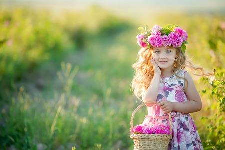 A little girl with beautiful long blond hair, dressed in a light dress and a wreath of real flowers on her head, in the garden of a tea rose.の写真素材