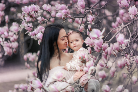 Cute Baby 6 month old Girl in Pink Outfit with Big Blue Eyes with Young Beautiful Mother at Spring, Pink Blooming Tree at the Background.の写真素材