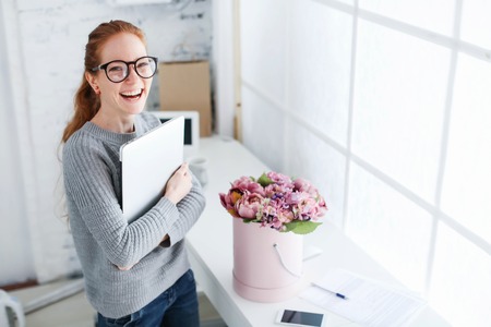 Young beautiful woman with red hair, wearing glasses, working in the office, uses a laptop and mobile phone.の写真素材