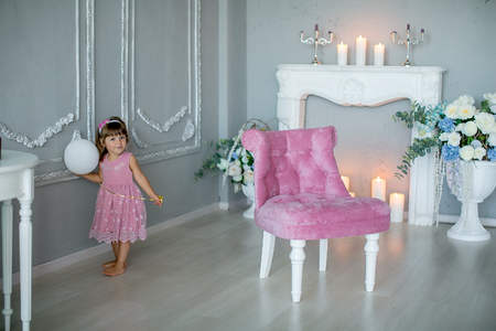 Smiling baby girl under 1 year old wearing stylish clothes sitting in vintage chair over white fireplace in room. Holiday season. Childhoodの写真素材