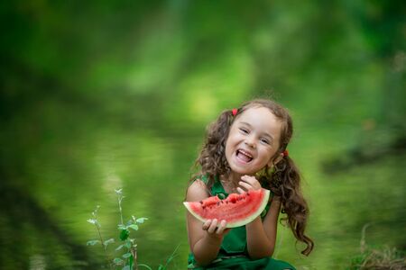 Cute girl sits on the banks of the river and eats a slice of fresh red watermelon.の写真素材