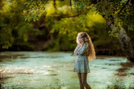 A young girl with long blond curly hair and a summer skirt poses on the Bank of the river in the spring green forest.の写真素材