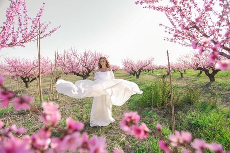 An adult woman in a beautiful white dress runs full length through a garden with trees in pink flowers.の写真素材