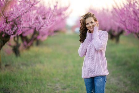 a stylish adult woman in a glamorous sweater near the branches of a tree covered with pink blossoms.の写真素材