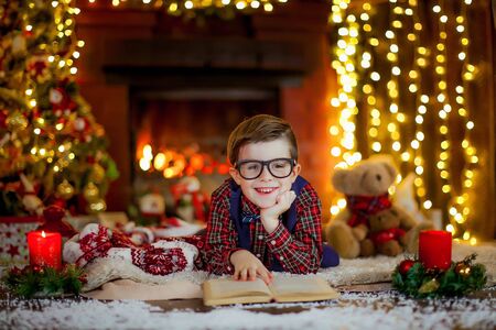 A boy of 5 years lies on his stomach against the background of the fireplace and reads a book. New Years Eve in a cozy home interior.の写真素材