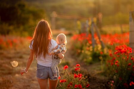 A woman walks with her back to the camera across a field of poppies and carries a child in her arms.