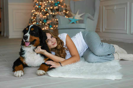 A beautiful woman with long hair lies on a rug against the background of a Christmas tree and next to her lies her big dogの写真素材