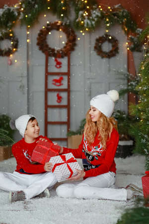 Mom and adult son sitting on the ground surrounded by artificial snow wearing hats white pants and a...