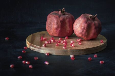 Two old ugly dried-up red pomegranates fruits with seeds on little wooden cutting board over dark background. Side view. horizontal orientation. Close up.の写真素材