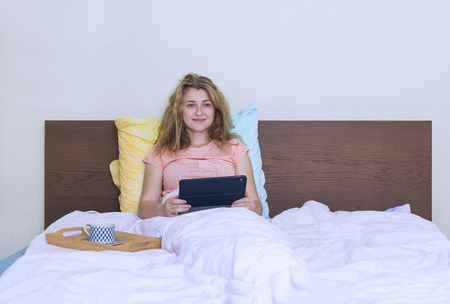 1 girl in bed under a blanket with a gadget and a Cup of coffee, young woman with curly hair smiling, restingの写真素材
