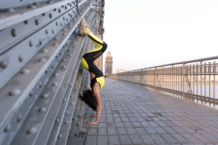 1 white athletic girl in black and yellow tracksuit standing on her hands upside down on the bridge...