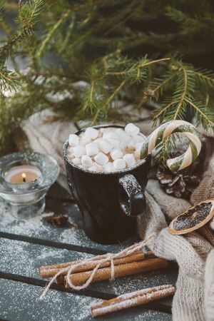 mug of hot coffee, chocolate with lots of marshmallows and candy, the cinnamon, the candle next to the knitted fabric on a background of green fir branches, winter hot drink, the Christmas spiritの写真素材
