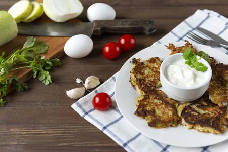 plate with zucchini pancakes with sour cream and herbs on a wooden background, knife, fork, fresh...