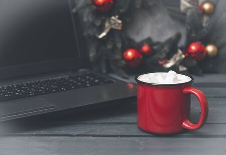 1 red mug of hot chocolate or coffee and marshmallows next to a spruce Christmas wreath with red...