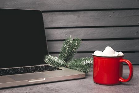 1 red mug with hot chocolate, cocoa and marshmallows next to laptop and fir branch on grey backgroundの写真素材