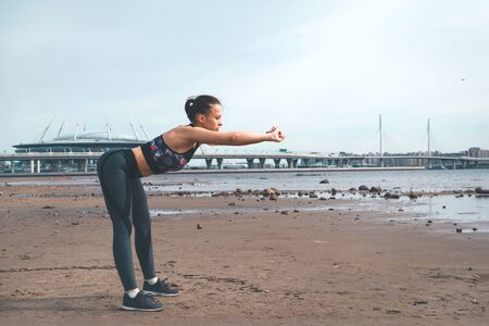 1 white slender European girl in sportswear leans forward on the beach against the city background...