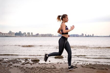 1 white slim athletic European girl in sportswear runs along the beach along the sea shore against...