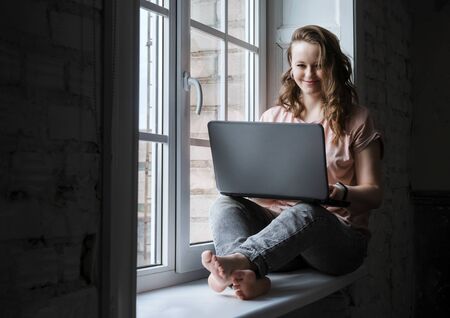 1 beautiful young European woman sitting on the windowsill near the window with a laptop and...