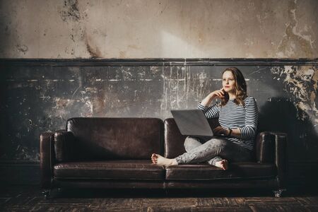 1 a white young woman in gray jeans and a vest with a laptop sits on a sofa against an old wall in the sunlightの写真素材