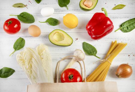 products are poured out of a paper bag, pasta, eggs, fruit, vegetables, lettuce, pepper, tomato, lemon , avocado, spinach, garlic, onion on a white wooden backgroundの写真素材