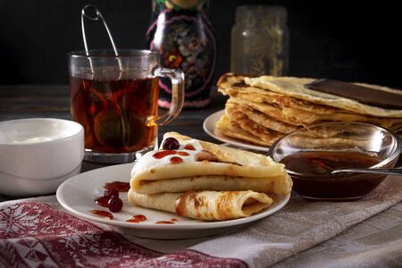 several pancakes with berries, sour cream and jam on a plate, a mug of black tea on a towel on a dark wooden backgroundの写真素材