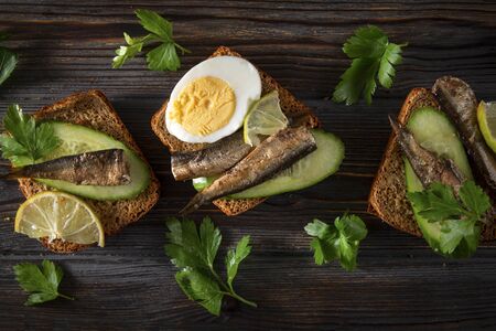 3 sandwiches with sprats, fresh cucumber, parsley, lime, rye bread  and boiled egg on a dark wooden background, top view, canned fish,の写真素材