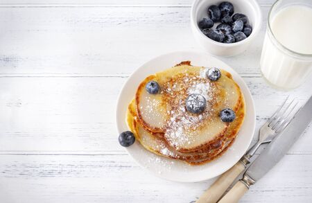 fried pancakes with  blueberries on a plate, a glass of milk, a fork, a knife on a white wooden background,の写真素材