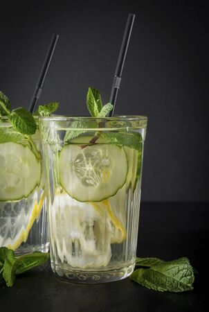 2 glasses of lemonade with yellow lemon, green mint , slices of fresh cucumber  and tubes on a black background, copy space, close upの写真素材