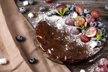 homemade chocolate cake with fresh berries, red grapes and blueberries, mint, coconut crumbs, powdered sugar and marshmallows on a beige towel on a wooden brown background, bakingの写真素材