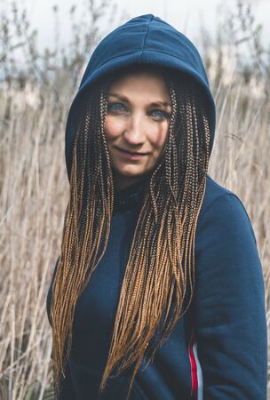 portrait 1 of a modern beautiful white woman with African pigtails in a dark dress with a hood on a background of dry grassの写真素材