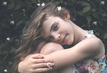 portrait of 1 white girl 10 years old with long hair close up on the background of greenery in nature, sitting, hugging her kneesの写真素材