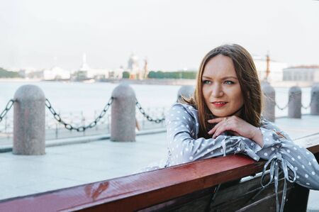 portrait of a white woman with red hair, a girl sitting on a bench on the embankment of the Neva river against the background of the city on a summer dayの写真素材