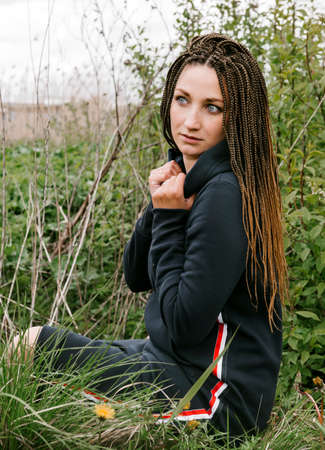 1 white young woman in Afro pigtails in a dark dress sitting on grass outdoors, portrait of a...