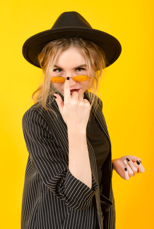 1 stylish young blonde woman in a black suit, hat and yellow glasses on a yellow backgroundの写真素材