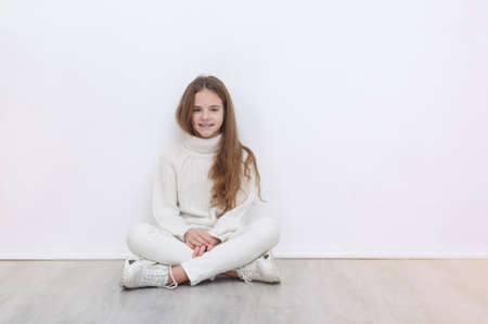 1 white girl 10 years old in a white sweater, jeans and sneakers sits on the floor against a white wall and smiles, teenagerの写真素材