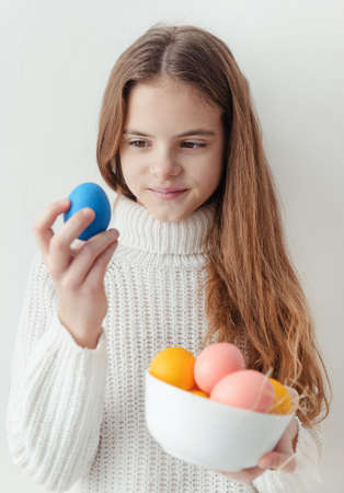 1 white cute European girl 10 years old with blue yellow and pink Easter eggs in a white sweater on a white wall backgroundの写真素材