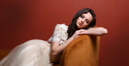 Beautiful white young woman in a white dress sitting on a sofa against a red wall backgroundの写真素材