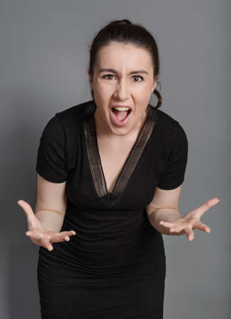 angry young woman in black dress gets angry and screams on gray background