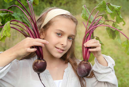 white girl of 10 years old holds 2 beets in her hands on a background of greeneryの写真素材